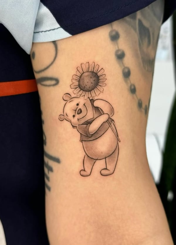 A black and grey fine line tattoo of Winnie the Pooh holding a sunflower on the upper arm, featuring delicate linework and shading — by otaybeatdown, orange, ca