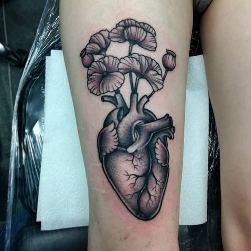 A bold black and grey tattoo of an anatomical heart with flowers on the thigh — by Samuel Connelly, los angeles