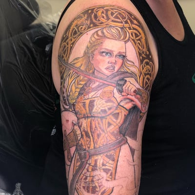 A colorful illustrative tattoo of a warrior woman with Celtic designs on the upper arm, featuring a sword and intricate patterns — by Sandra Price, los angeles