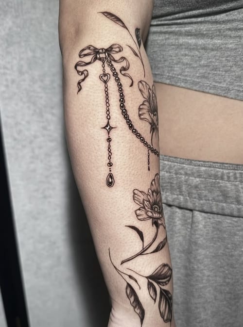 A delicate black and grey fine line tattoo on the forearm featuring floral designs and a dangling chain with a bow — by Shelby Bianco, los angeles