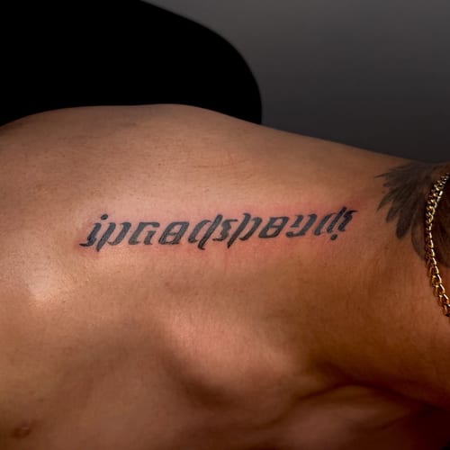 A bold black script lettering tattoo on the shoulder with a clean and striking design — by NJ TATTOOZ, los angeles