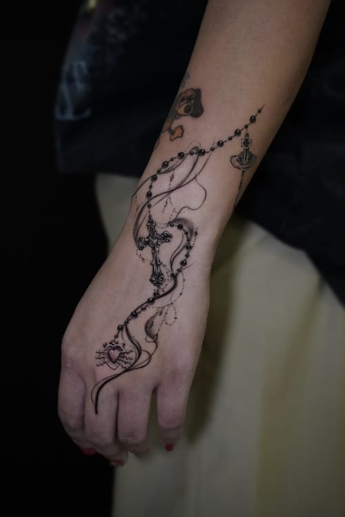 A fine line black and grey tattoo on the hand featuring a rosary with a cross and heart, elegantly wrapping around the wrist and fingers — by Jian Liang, los angeles