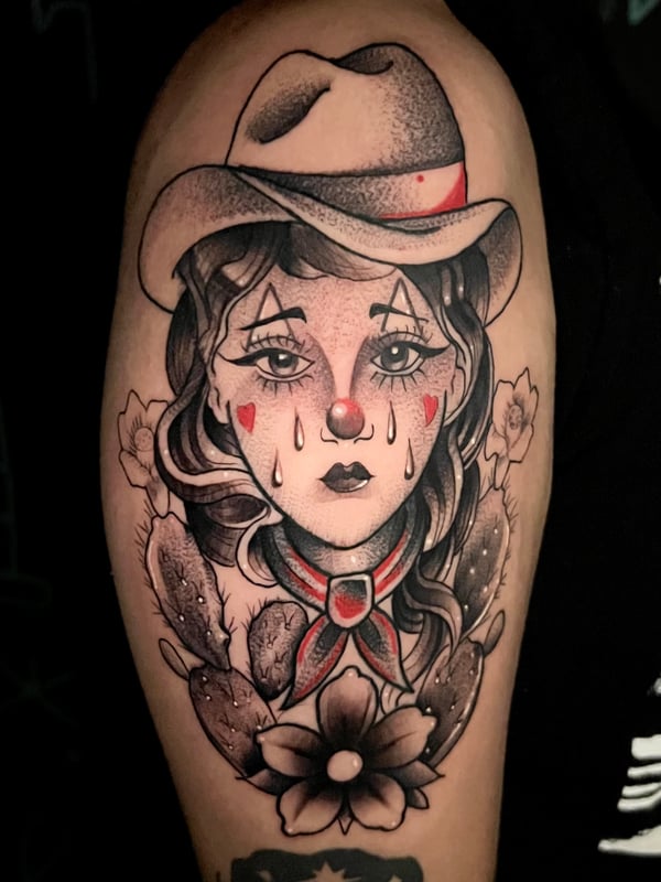 A Neo-Traditional tattoo of a crying woman with a clown theme, wearing a hat, surrounded by flowers on the upper arm. The design uses black and grey shading with red accents — by Josue Bravo, los a…