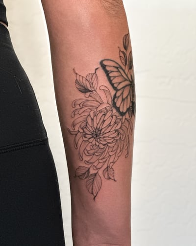 A delicate black and grey fine line tattoo of a butterfly and flower on the forearm — by Melanie Benoit, los angeles
