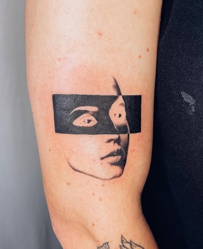 A minimalist blackwork tattoo of a partially obscured face on the upper arm, featuring a bold black rectangle across the eyes — by Level tattoo, los angeles