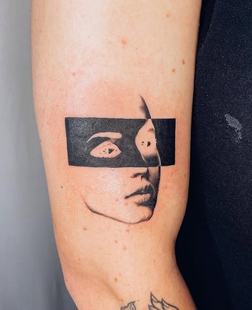 A minimalist blackwork tattoo of a partially obscured face on the upper arm, featuring a bold black rectangle across the eyes — by Level tattoo, los angeles