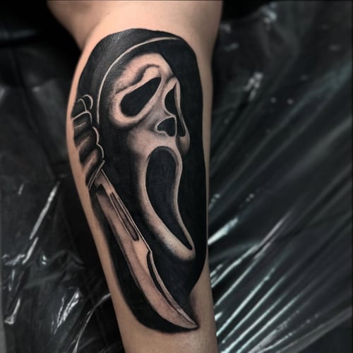 A black and grey Ghostface mask with a knife tattoo on the leg, featuring bold blackwork style — by Maddie Ramirez, los angeles