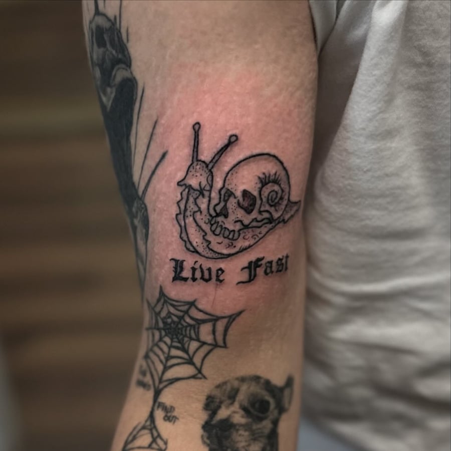 A bold blackwork tattoo of a snail with a skull shell and 'Live Fast' text on the arm — by Maddie Ramirez, los angeles