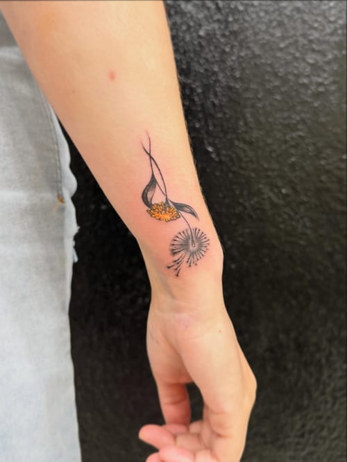 A delicate minimalist dandelion tattoo on the forearm with fine line work and subtle color accents — by Kiera Garvin, los angeles