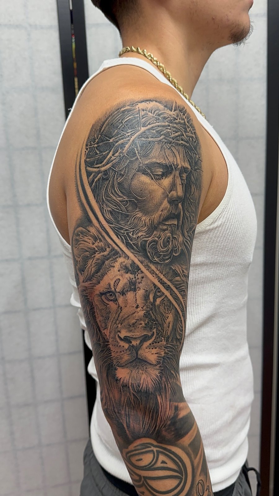 A detailed black and grey realism tattoo of Jesus Christ and a lion on the upper arm, showcasing intricate shading and texture — by Humilde tattoos, los angeles