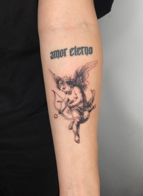 A black and grey fine line tattoo of a cherubic angel with a bow and arrow on the forearm, accompanied by script text above — by ashynucklestattoo, los angeles