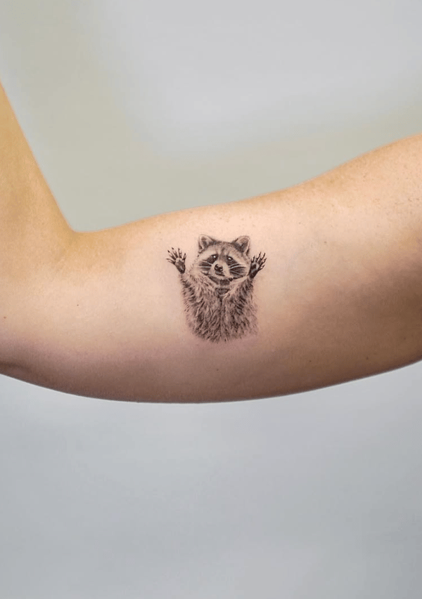 A small black and grey realistic tattoo of a raccoon on the inner arm, featuring detailed fur and a playful expression — by Jian Liang, los angeles