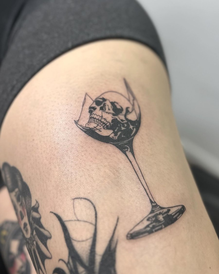 A black and grey tattoo of a skull inside a broken wine glass on the thigh, done in blackwork and fine line style — by Maddie Ramirez, los angeles