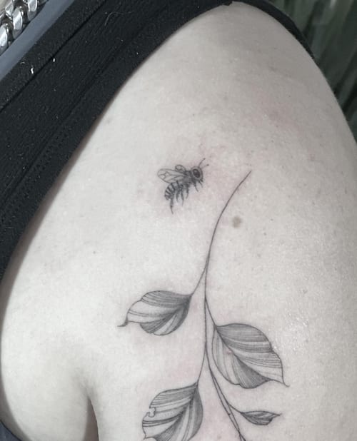 A delicate black and grey tattoo of a bee and leaves on the upper arm, done in a fine line minimalist style — by Melanie Benoit, los angeles