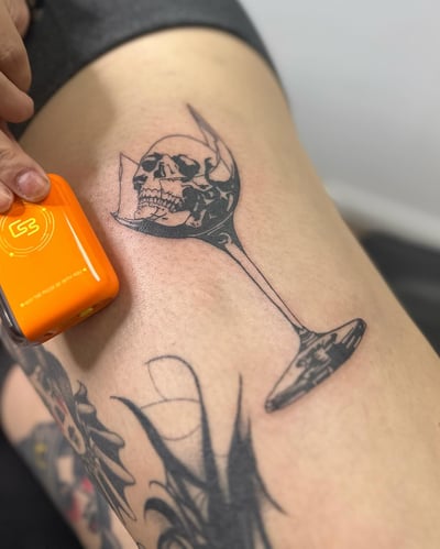 A black and grey tattoo of a skull inside a broken wine glass on the thigh, done in blackwork and fine line style — by Maddie Ramirez, los angeles