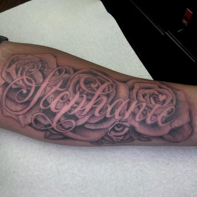 A bold black and grey script lettering tattoo on the forearm featuring roses and intricate shading — by Sandra Price, los angeles