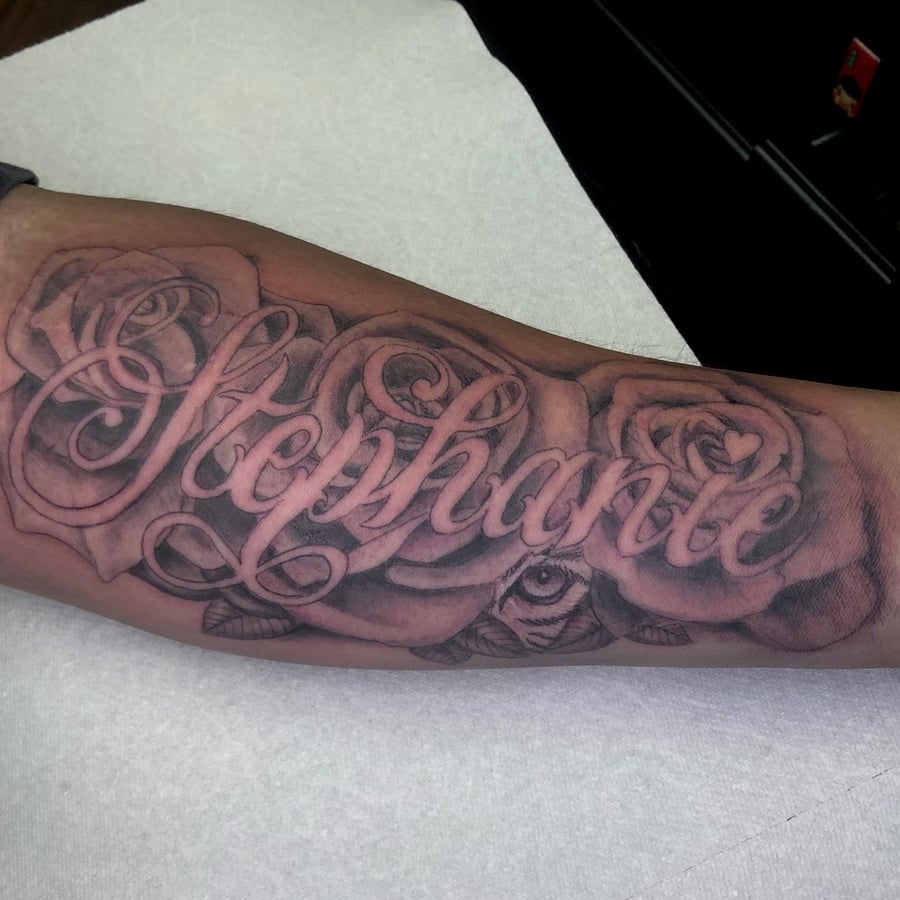 A bold black and grey script lettering tattoo on the forearm featuring roses and intricate shading — by Sandra Price, los angeles