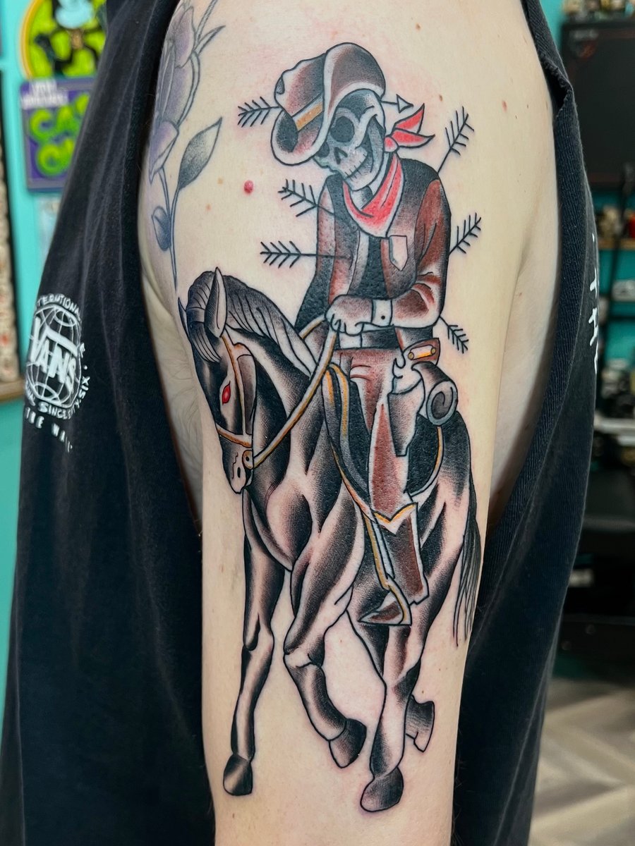 A neo-traditional tattoo of a skeleton cowboy riding a horse on the upper arm, featuring black and grey shading with subtle red accents — by Josue Bravo, los angeles