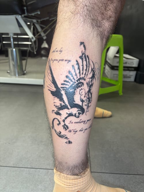 A black and grey tattoo on the calf featuring a stylized bird in flight with accompanying script, rendered in a bold blackwork style — by Hannah , los angeles
