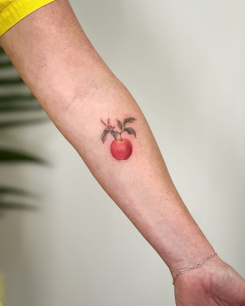 A realistic color tattoo of an apple with leaves on the forearm, featuring fine line details and vibrant colors — by Melanie Benoit, los angeles