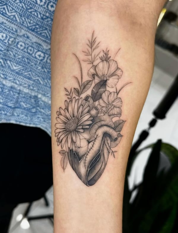 A black and grey fine line tattoo on the forearm depicting an anatomical heart intertwined with various flowers, showcasing intricate details and delicate shading — by otaybeatdown, orange, ca