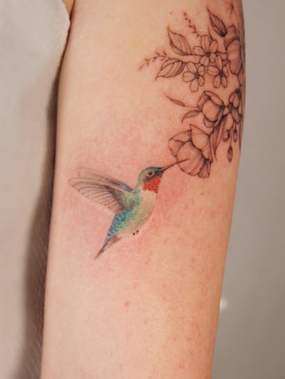 A colorful hummingbird and fine line floral tattoo on the arm, featuring delicate and realistic details — by Melanie Benoit, los angeles