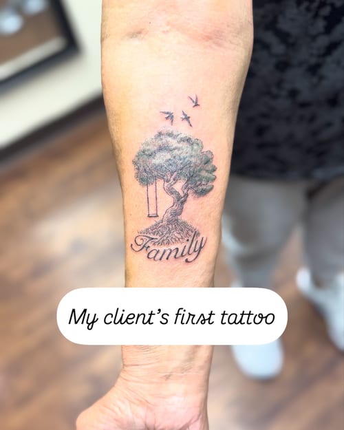 A minimalist black and grey tattoo of a tree with a swing and birds, featuring the word 'Family' on the forearm — by Sandra Price, los angeles