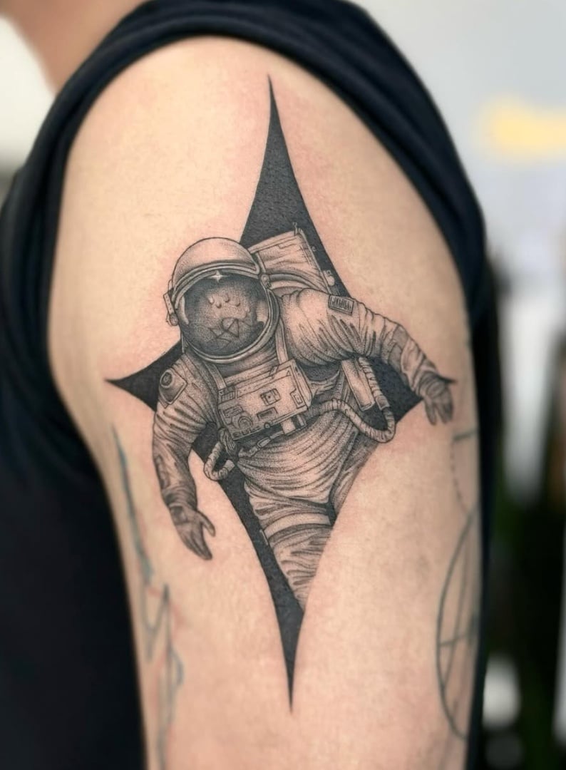 A black and grey realism tattoo of an astronaut with an animal face on the upper arm, featuring detailed shading and a star-shaped background — by otaybeatdown, los angeles