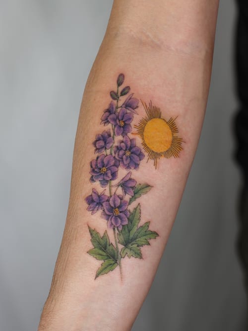A delicate color tattoo of purple flowers and a sun on the forearm, featuring a botanical style — by Melanie Benoit, los angeles