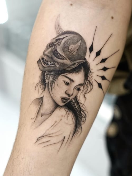 A black and grey tattoo on the forearm depicting a realistic portrait of a woman wearing a traditional Japanese mask, with detailed shading and fine lines — by otaybeatdown, los angeles