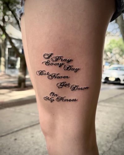 A black and grey lettering tattoo on the thigh featuring a quote in elegant script — by emily, los angeles
