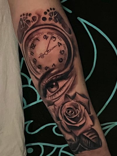A black and grey realistic tattoo on the forearm featuring a detailed eye, a clock with Roman numerals, and a rose, all intricately shaded — by Josue Bravo, los angeles