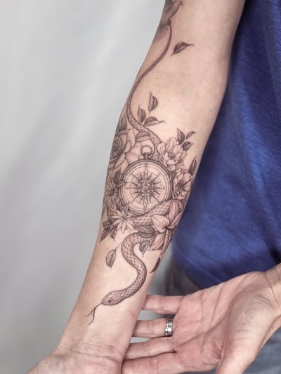 A delicate black and grey tattoo of a snake intertwined with a compass and flowers on the forearm, featuring fine line and blackwork styles — by Melanie Benoit, los angeles