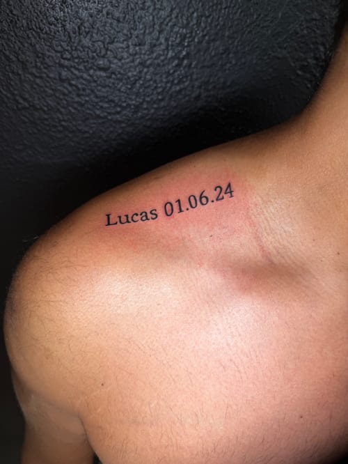 A minimal black script tattoo on the shoulder featuring a name and date in simple lettering — by Kiera Garvin, los angeles