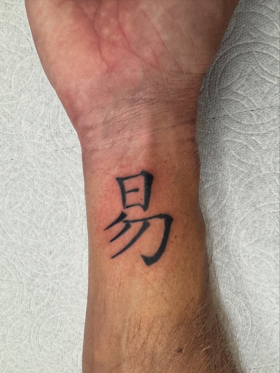 A minimalist black ink Chinese character tattoo on the wrist, featuring simple and bold lettering — by Sandra Price, los angeles