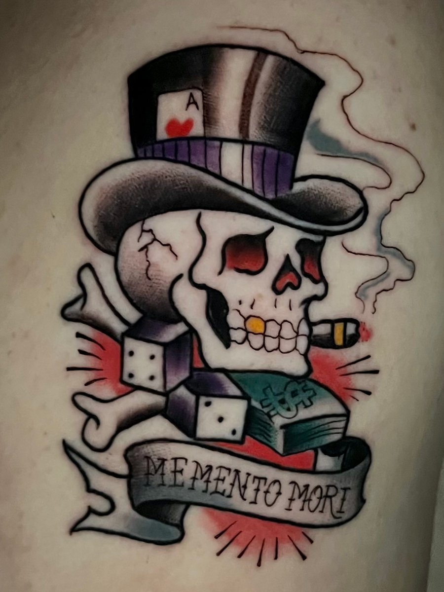 A colorful neo-traditional tattoo on the arm featuring a skull with a top hat, dice, and a banner with 'Memento Mori' script, accented with red and green highlights — by Josue Bravo, los angeles
