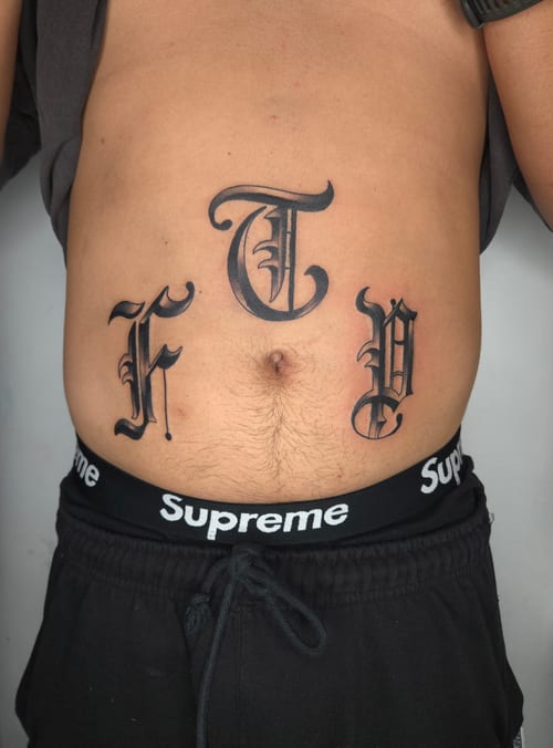 Bold black and grey lettering tattoo on the stomach, featuring ornate script letters — by ashynucklestattoo, los angeles