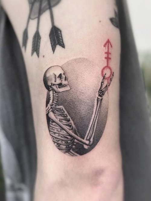 An illustrative tattoo of a skeleton holding a red symbol, with black and grey shading on the inner arm — by Jian Liang, los angeles