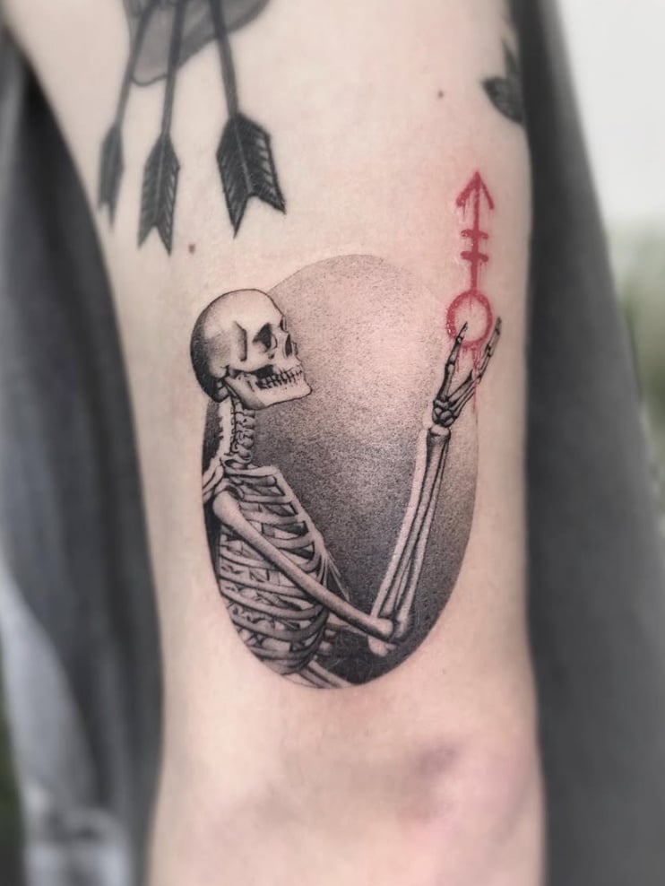 An illustrative tattoo of a skeleton holding a red symbol, with black and grey shading on the inner arm — by Jian Liang, los angeles