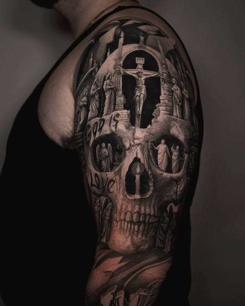 A detailed black and grey tattoo on the upper arm depicting a religious scene with Jesus Christ on the cross, integrated into a skull design — by Vladimir Kozachenko, los angeles