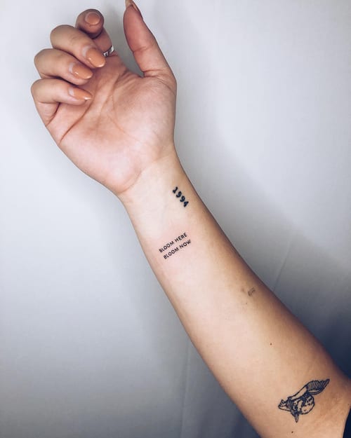 A minimalist blackwork tattoo on the wrist featuring the number '1994' and the quote 'Bloom Here Bloom Now' in simple script — by Level tattoo, los angeles