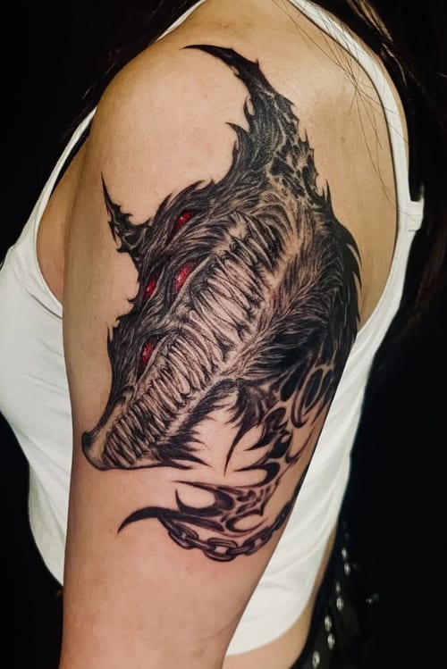 A black and grey tattoo of a demonic face with sharp teeth and red eyes on the upper arm, featuring bold blackwork and dark art elements — by zitronxus, los angeles