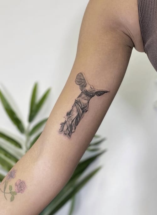 A black and grey realism tattoo of the Winged Victory of Samothrace on the upper arm, showcasing detailed shading and classical sculpture style — by Jian Liang, los angeles