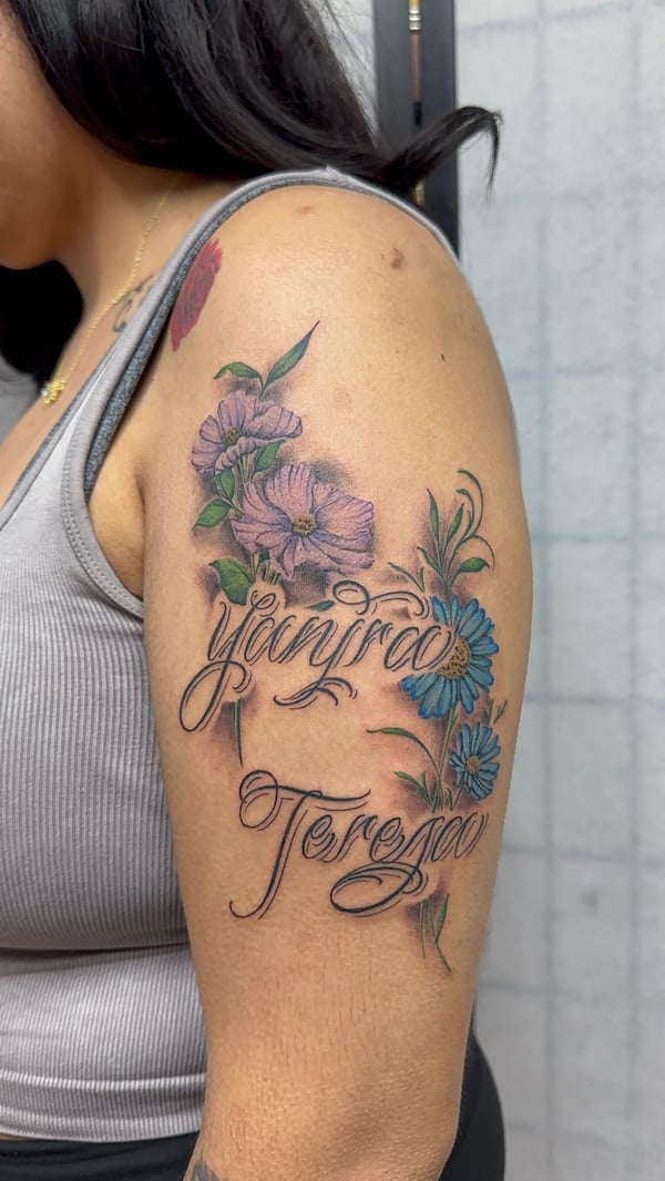 A colorful floral tattoo with script lettering on the upper arm, featuring purple and blue flowers with green leaves — by Humilde tattoos, los angeles