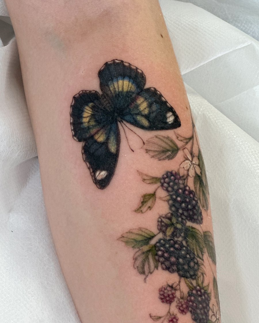 A realistic color tattoo of a butterfly and blackberries on the forearm, featuring delicate botanical details — by Melanie Benoit, los angeles