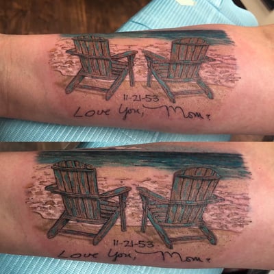 A realistic color tattoo of two beach chairs on a sandy shore with ocean waves, accompanied by a memorial script on the forearm — by Sandra Price, los angeles
