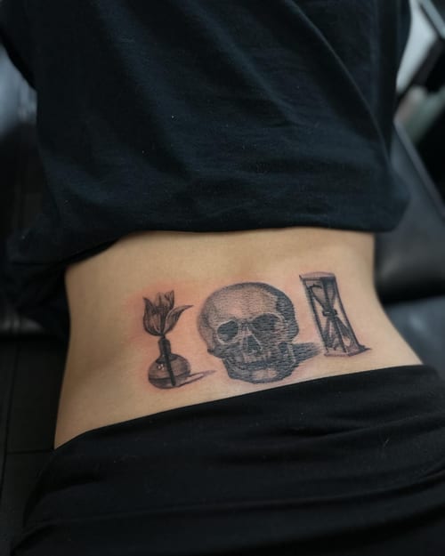 A black and grey tattoo of a skull with an hourglass and flower on the lower back — by Maddie Ramirez, los angeles