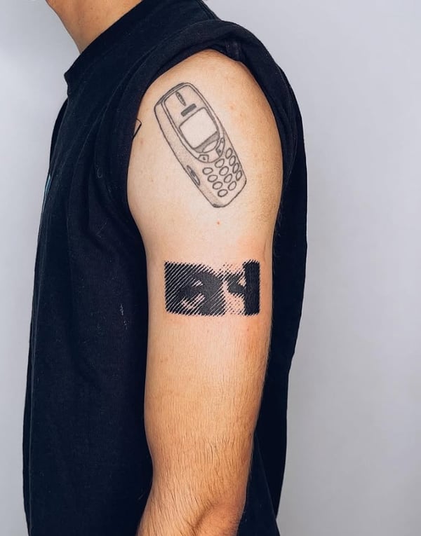 A minimalist black and grey tattoo of a vintage mobile phone and a pixelated design on the upper arm — by Level tattoo, los angeles