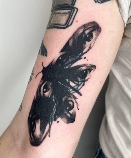 A surreal black and grey butterfly tattoo on the arm, with eyes incorporated into the wings, creating a unique and striking design — by Level tattoo, los angeles