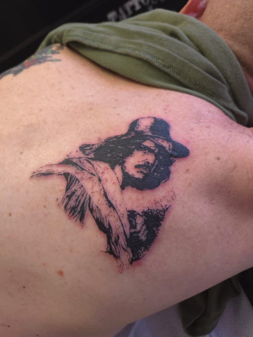 A black and grey portrait tattoo on the back, depicting a person with a hat and detailed shading — by ashynucklestattoo, los angeles